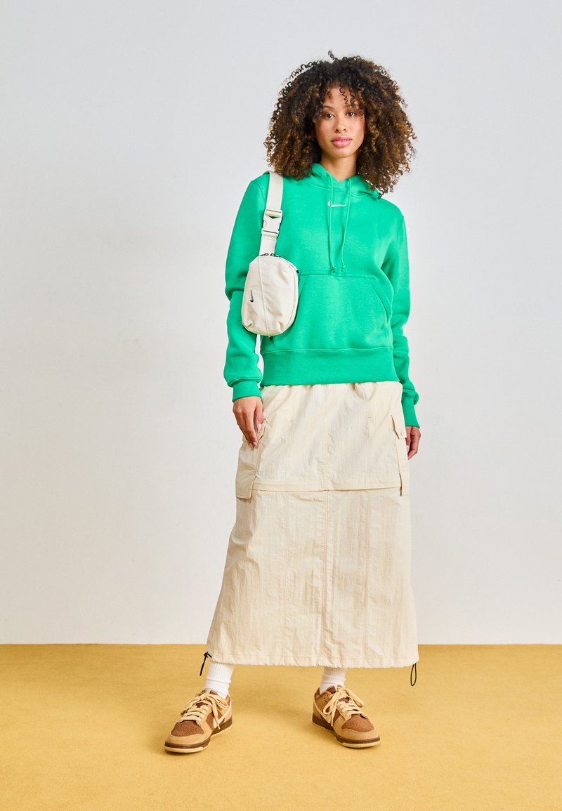 Groene hoodie, beige rok met zakken en bruine sneakers. Krullend haar, crèmekleurige schoudertas en witte sokken maken de outfit compleet.