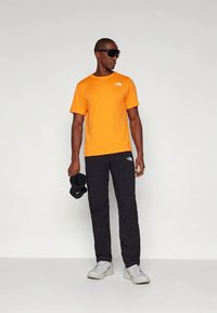 Oranžna majica s kratkimi rokavi z logotipom, kombinirana s črnimi cargo hlačami in belimi supergami. Dodatki vključujejo črno kapo, ki jo dr�žijo v roki.