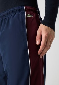 Jūras zila un bordo sporta šorti ar elastīgu jostu, baltām sānu svītrām un zaļu izšuvumu logotipu kreisajā pusē.