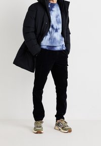 Czarna pikowana kurtka, niebieski sweter tie-dye, czarne sztruksowe spodnie oraz beżowo-zielone sneakersy z szarymi akcentami. Casualowa, warstwowa stylizacja.