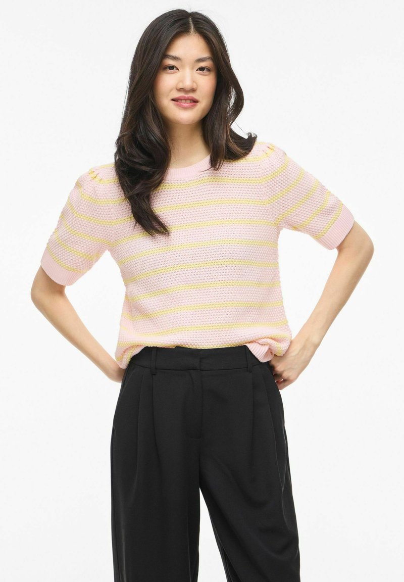 Jeune femme aux longs cheveux bruns portant un pull à manches courtes rayé rose et jaune et un pantalon taille haute noir, les mains sur les hanches.