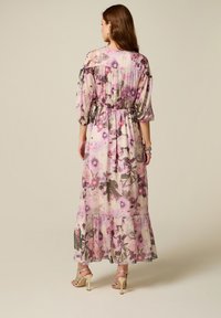 Mujer con un vestido largo y transparente con flores rosas y moradas, mangas 3/4, cintura fruncida y bajo con volantes, combinado con sandalias de tacón alto.
