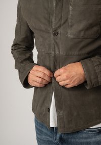 Dunkelolivgrüne Lederjacke mit Knopfleiste, zwei Brusttaschen und sichtbaren Nähten. Lässiger Schnitt mit leicht strukturiertem Oberflächen.