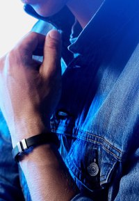 Svart lærarmbånd med to hvite detaljer, båret på en hånd. Personen har på seg en denimjakke som viser teksturert stoff og en knappdetalj.