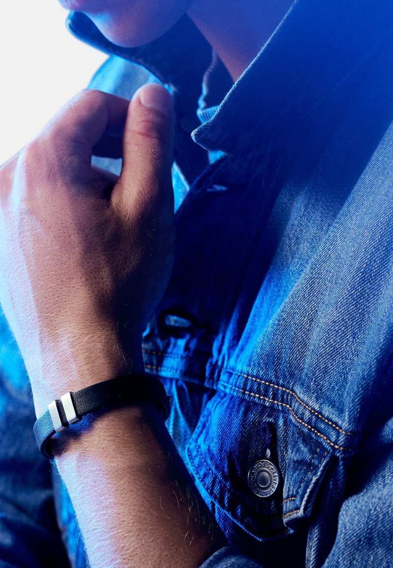 Svart lærarmbånd med to hvite detaljer, båret på en hånd. Personen har på seg en denimjakke som viser teksturert stoff og en knappdetalj.