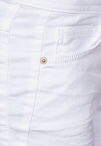 Witte denimstof met een textuur heeft een zijzak en een metalen studaccent. De stiksels zijn zichtbaar en netjes.