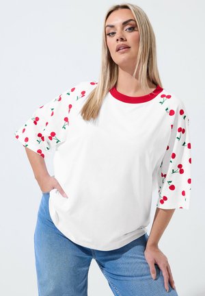 Woman wearing a white T-shirt with red cherry prints on sleeves and red neckline, paired with blue jeans, posing with one hand on hip.
