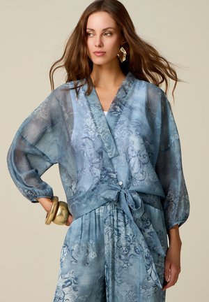 Femme aux longs cheveux bruns portant un chemisier portefeuille bleu transparent à motifs floraux et un pantalon assorti, accessoirisée de larges bracelets et boucles d'oreilles en or.