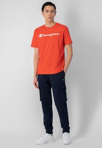 Camiseta de algodón roja con el logo "Champion" en blanco, combinada con pantalones de chándal cargo en color marino que tienen bolsillos laterales, y zapatillas blancas.