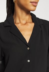 Chemise noire à col avec un tissu texturé, présentant un décolleté en V et deux boutons de couleur claire à l'avant.