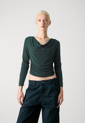 Green long-sleeve top with a cowl neck, featuring gathered fabric and a fitted design, paired with high-waisted dark trousers.