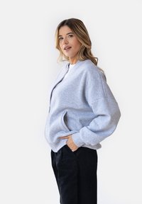 Jasnoszara bluza zapinana na zamek z oversize'owymi rękawami i przednimi kieszeniami. Tkanina wydaje się miękka o gładkiej fakturze. Nosi się ją na białej bluzce.