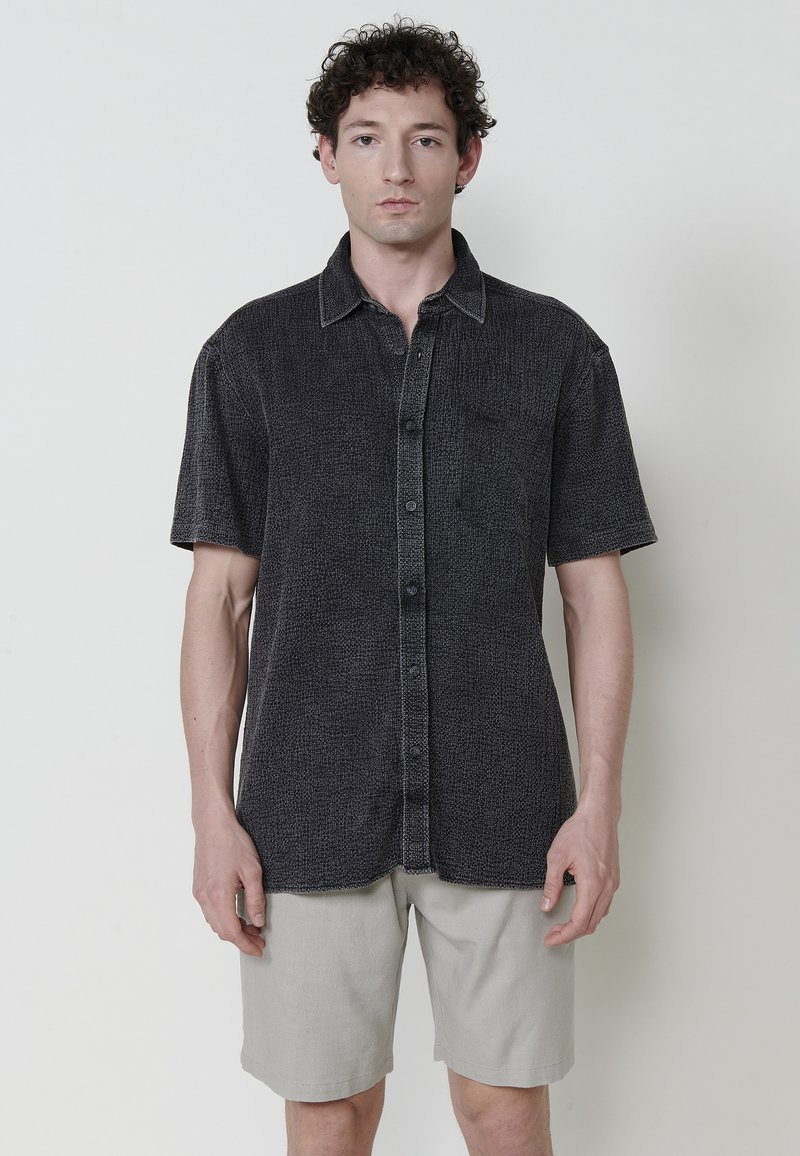 Camisa de manga corta negra con un patrón texturizado, con botones en el frente y un cuello sutil, combinada con pantalones cortos beige claros.