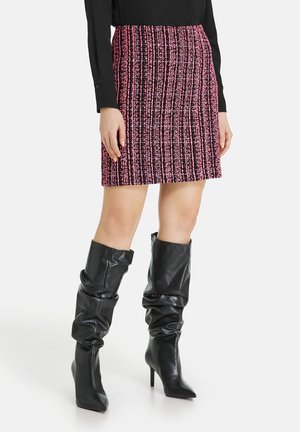 Vrouw draagt een roze en zwarte textuur minirok, zwarte top met lange mouwen en zwarte slordige hoge hakken knie laarzen.