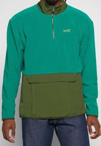 Chaqueta de pullover verde con cuello alto, cierre de cremallera y un panel inferior verde adicional. Presenta un bolsillo frontal y detalle del logo de Levi's.