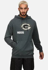Kolgrå hoodie med en stor oval "G"-logotyp och texten "PACKERS". Tillverkad av mjukt material med en framficka och justerbar huva.