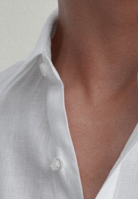 Hals en bovenste borst van een persoon die een ongebonden wit linnen overhemd draagt met zichtbare kraag en knopen.