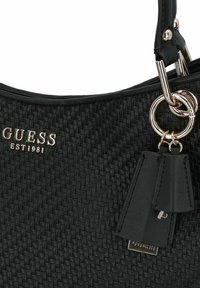 Borsa a mano nera intrecciata con tessuto texture, hardware in argento e un'etichetta decorativa. Presenta il logo del marchio "GUESS EST1981" ben visibile.