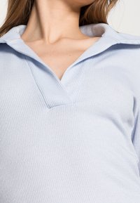 Haut côtelé bleu clair avec un col et une découpe en V peu profonde. Le tissu semble doux, offrant une finition texturée.