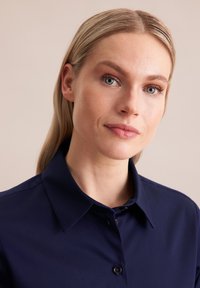 Granatowa koszula z długim rękawem, z ostrym kołnierzykiem, gładką teksturą i pojedynczym rzędem guzików. Zbliżenie na dekolt i ramiona.