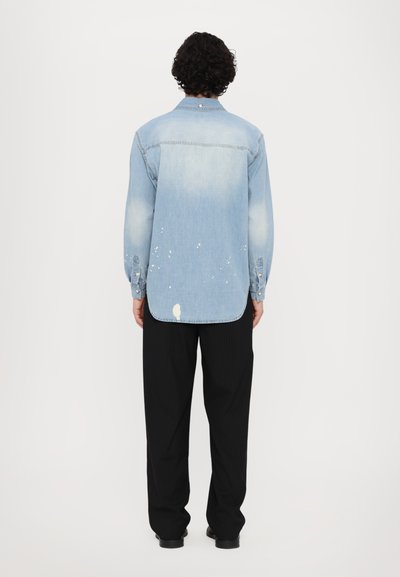 Camicia di jeans azzurro chiaro con un lavaggio sbiadito, orlo leggermente sfrangiato e dettagli di schizzi di vernice bianca. Maniche lunghe con polsini abbottonati.