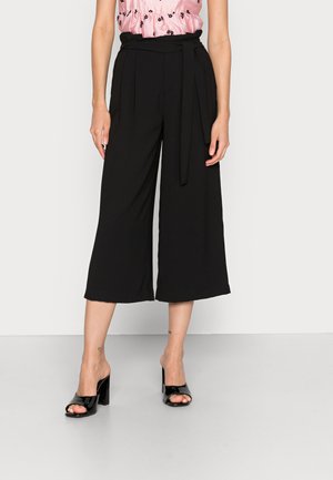 Culottes largas pretas feitas de tecido suave, com uma cintura cingida. Combinadas com mules pretas de salto aberto. Modelo posa contra um fundo simples.