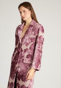 Abrigo largo con estampado floral en tonos rosas y beiges, con cuello de chal, dos bolsillos frontales y una tela texturizada.