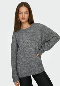 Szary, oversize sweter z dzianiny z opadającymi ramionami, okrągłym dekoltem i fakturowanym materiałem. Stylizowany z czarnymi spodniami i długimi kolczykami.