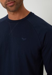 Granatowa koszulka z długim rękawem z okrągłym dekoltem, z dyskretnym haftowanym logo i kontrastowym przeszyciem przy dekolcie oraz szwach ramion.