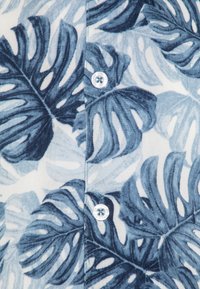 Camicetta a maniche corte con un motivo di foglie botaniche blu su uno sfondo chiaro. Include un bottone bianco e una texture sottile.