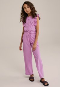 Růžový texturovaný overal s volánkovými ramenními akcenty, předním zapínáním na knoflíky a nastavitelným páskem v pase; doplněný černými tkanými sandály.