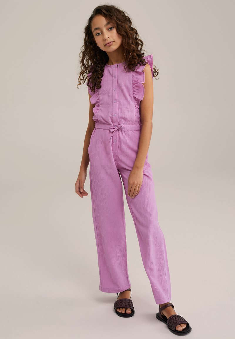 Růžový texturovaný overal s volánkovými ramenními akcenty, předním zapínáním na knoflíky a nastavitelným páskem v pase; doplněný černými tkanými sandály.