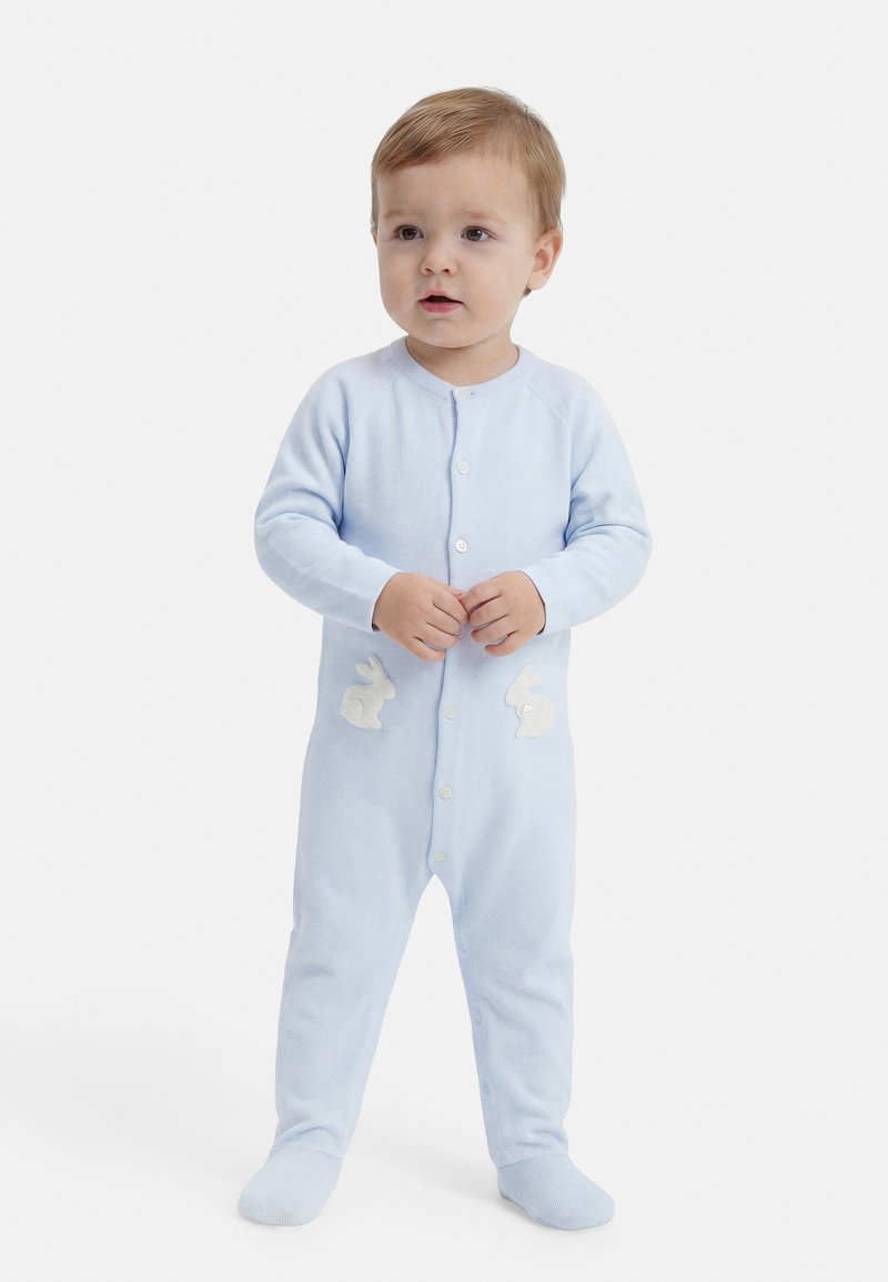 Νήπιο που στέκεται με ένα ανοιχτό μπλε φορμάκι με κουμπιά στο μπροστινό μέρος και λευκές σχεδιασμένες κουνελάκια στη κάθε πλευρά των γοφών, πάνω σε απλό λευκό φόντο.