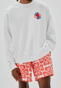 Witte sweatshirt met een gekleurd "GUESS BALDWIN" logo op de borst, gecombineerd met rode shorts met een hartpatroon en witte tekst.