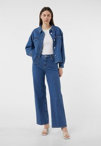 Dżinsowa kurtka i spodnie z wysokim stanem w średnim niebieskim kolorze. Kurtka posiada dwie kieszenie na piersi i guziki; spodnie mają luźny, rozszerzany krój oraz postrzępiony dół.