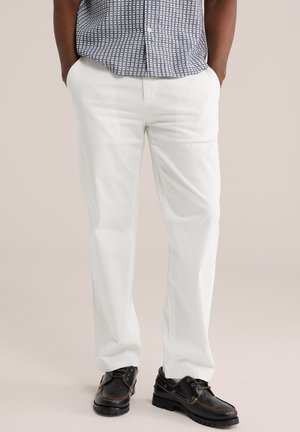 Uomo che indossa pantaloni bianchi a gamba dritta, camicia a maniche corte fantasia infilata dentro e scarpe nere stringate, in piedi con le mani in tasca.