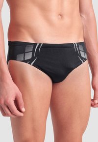 Männlicher Oberkörper in schwarzen Badebrieftaschen mit weißen geometrischen Mustern an den Hüften, stehend vor einem schlichten hellen Hintergrund.
