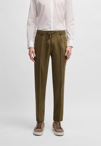 Olive green tapered pants with a drawstring waistband, made of smooth fabric. Paired with light brown textured sneakers and a white shirt.
