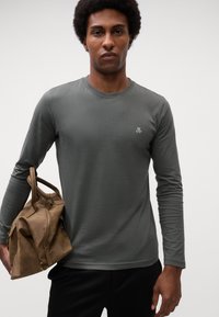 Camicia a maniche lunghe di colore verde oliva realizzata in un tessuto morbido, con scollo rotondo e un piccolo logo bianco sul petto. Una borsa beige è tenuta nella mano sinistra.