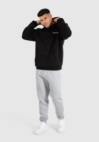 Sweat-shirt à capuche noire avec une poche avant et un logo, associé à un pantalon de survêtement gris et des baskets blanches. Le modèle ajuste sa capuche, se tenant sur un fond gris.