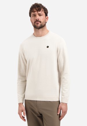 Homme aux cheveux courts et bouclés et à la barbe, portant un pull crème à manches longues et un pantalon marron, debout devant un fond clair uni.