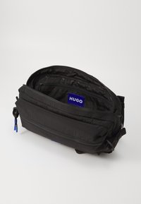 Sort nylon crossbody taske med flere lynlåslommer, der har et blåt logo og kontrasterende lynlåsdragter. Tekstureret overflade.