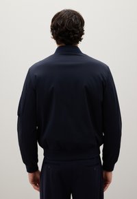 Giacca bomber blu navy con una texture liscia, colletto e polsini a coste e vita elastica. Presenta dettagli di cucitura sulle maniche.