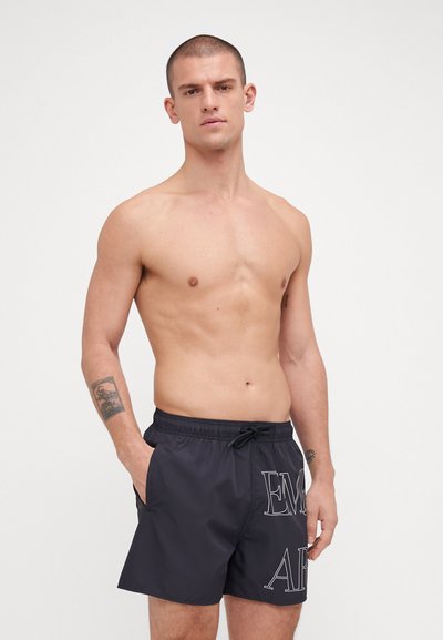 Shorts de bain noirs avec taille à cordon, poches latérales et grandes lettres blanches "EM" et "AR" sur la cuisse gauche. Matériau lisse et léger.