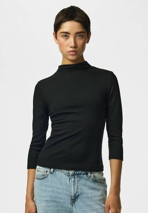 Ung person med kort mørkt hår iført en sort langærmet top og lyseblå jeans, poserer mod en ensfarvet hvid baggrund.