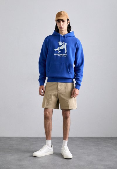 Felpa blu con una grafica e scritta bianche, abbinata a pantaloni beige e sneakers bianche. Il tessuto appare morbido con una vestibilità rilassata.