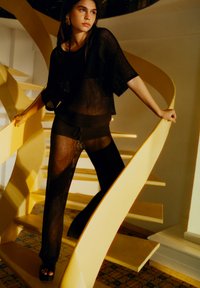 Černý síťovaný dvoudílný outfit s volnou blůzou a elastickými kalhotami, který má strukturovaný vzor. Modelka pózuje na žlutém točitém schodišti.