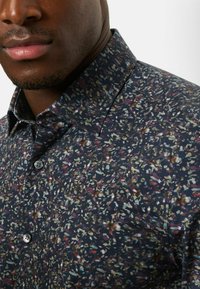 Knopfhemd in Dunkelblau mit floralem Muster in Grün-, Rosa- und Beigetönen. Weicher Stoff, spitzer Kragen und einzelne Knöpfe an den Manschetten.