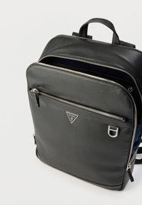Sac à dos Guess en cuir noir avec fermetures éclair argentées, poche avant zippée, poignée supérieure et plaque logo triangulaire, compartiment principal partiellement ouvert.