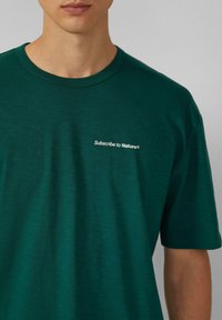 Olijfgroene katoenen T-shirt met een ronde halslijn en korte mouwen. Klein geborduurd "Subscribe to Nature®"-tekst op de voorkant.